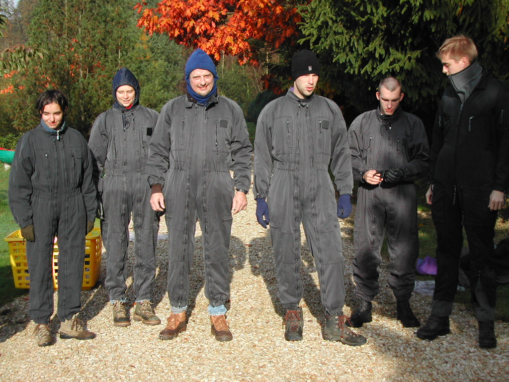 A group of six people wearing matching dark jumpsuits stands outdoors on a gravel path, dressed for an activity.