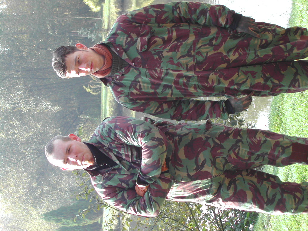Two people in camouflage clothing stand outdoors near water, with one crossing their arms and the other clasping their hands.