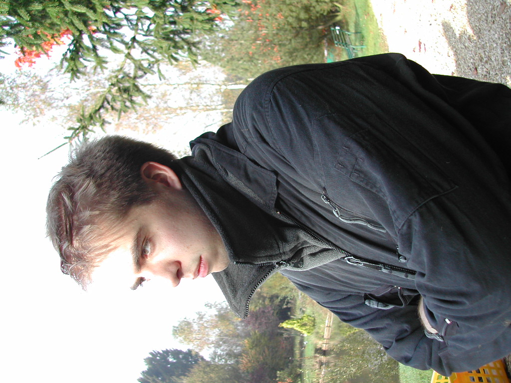 A person in a black jacket sits outdoors, looking to the side with a serious expression.