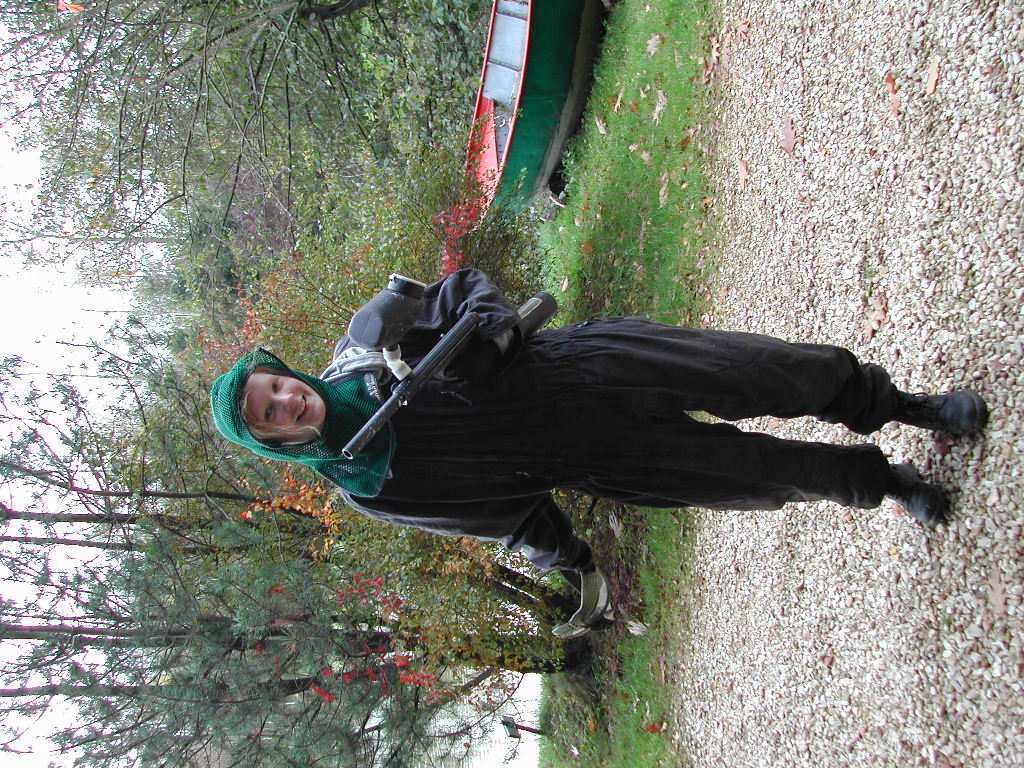 A person in black overalls and a green hood holds a paintball gun while standing on a gravel path.
