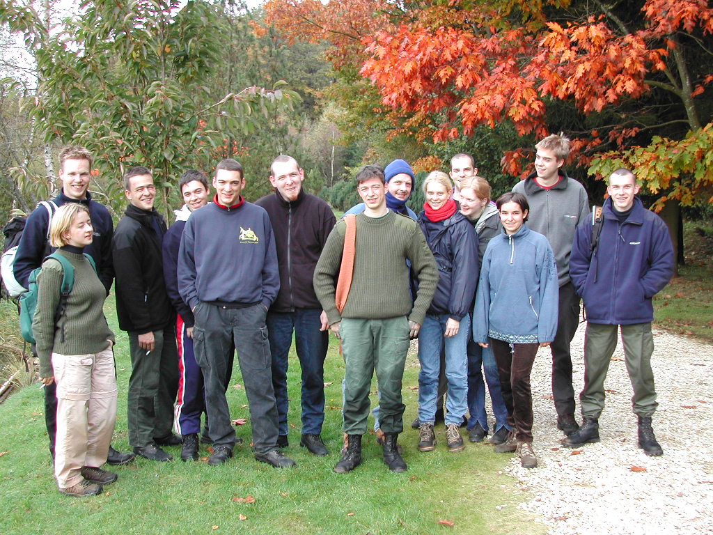 A group of people in outdoor clothing stand together, smiling, during a survival activity in a natural setting.