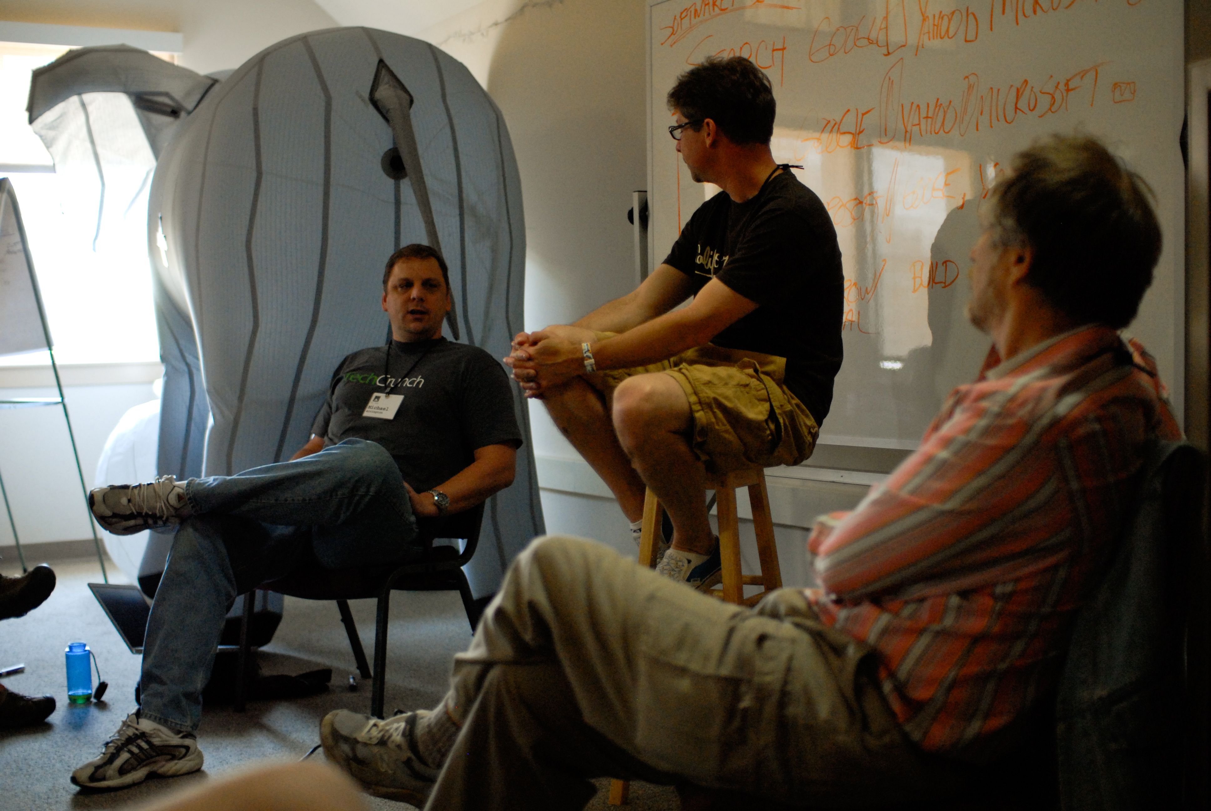 Two men engage in a debate about search engines while a moderator and others listen in a discussion setting.
