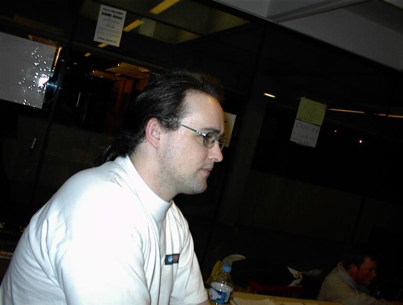 A man in a white t-shirt and glasses sits at a table, looking to the side.