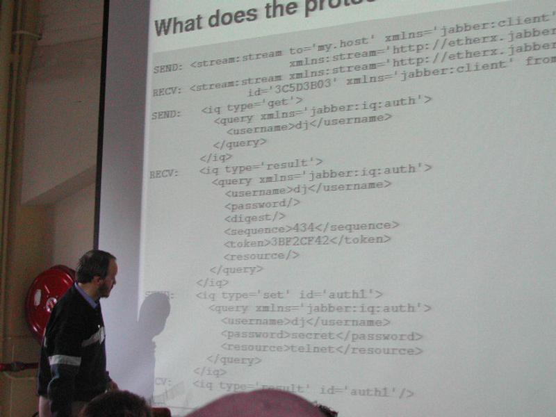A presenter stands in front of a projected slide displaying Jabber protocol code at a Fosdem 2002 event.