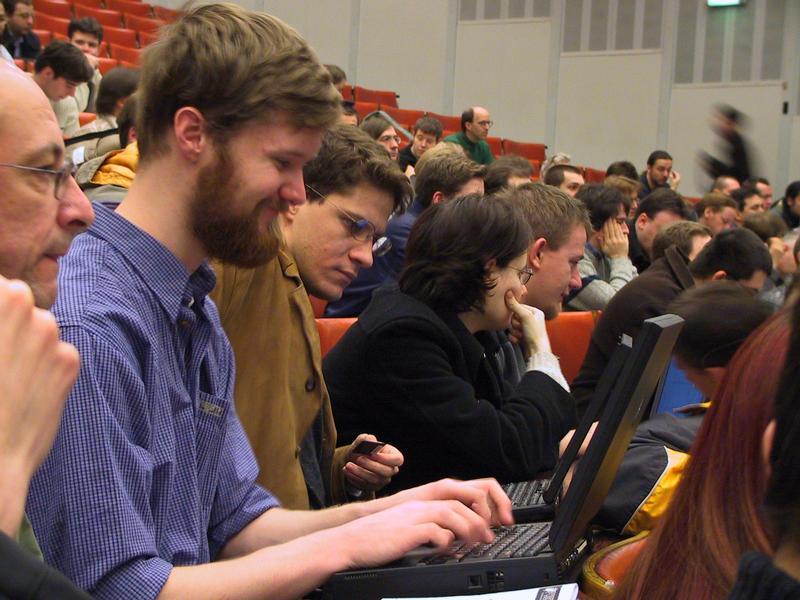 Two people in the foreground are using laptops in a crowded conference hall with attendees listening and taking notes.
