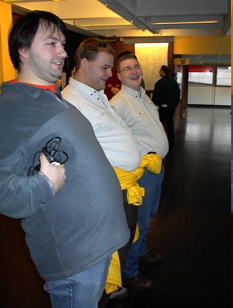 Three men stand in a row, leaning back and pushing out their stomachs in a playful pose indoors.