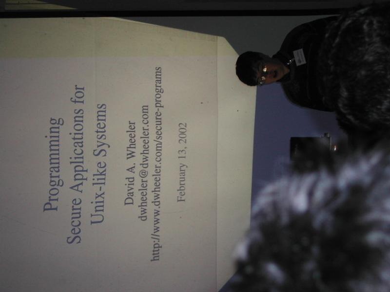 A speaker presents a talk on secure programming for Unix-like systems at Fosdem 2002, with a projected slide.