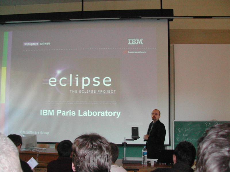 A presenter in a suit stands beside a projector screen displaying an "Eclipse" software slide at a conference.