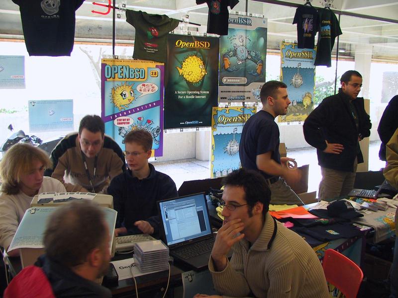 People gathered around a booth with OpenBSD posters, discussing and working on laptops at a technology event.
