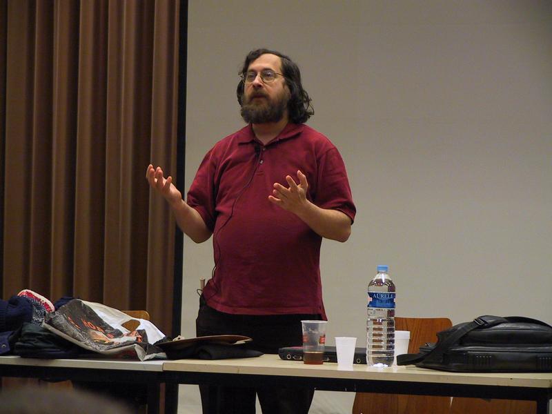 A man with a beard and glasses, wearing a red shirt, speaks with hand gestures in front of an audience.