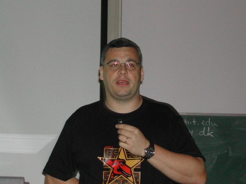 A man wearing glasses and a black T-shirt speaks into a microphone during a presentation at Fosdem 2002.