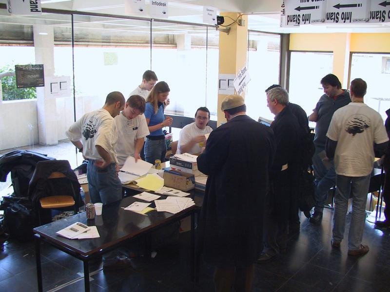 A group of people gathers around an information desk, engaging in discussions and handling papers at a conference.