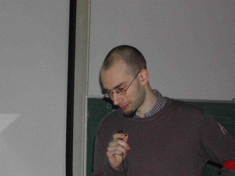 A person wearing glasses and a sweater holds a microphone while speaking at a Fosdem 2002 event.