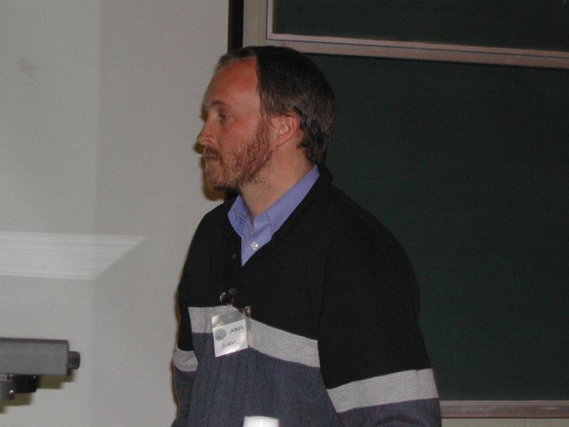 A person wearing a name badge stands in front of a chalkboard, appearing to give a presentation or lecture.