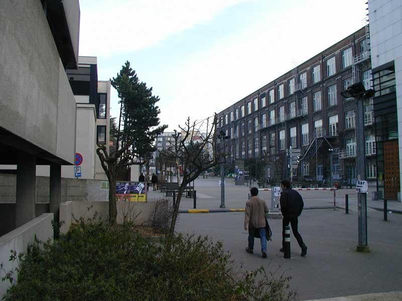 Two people walk along a paved path near buildings with posters and signs in an urban setting.