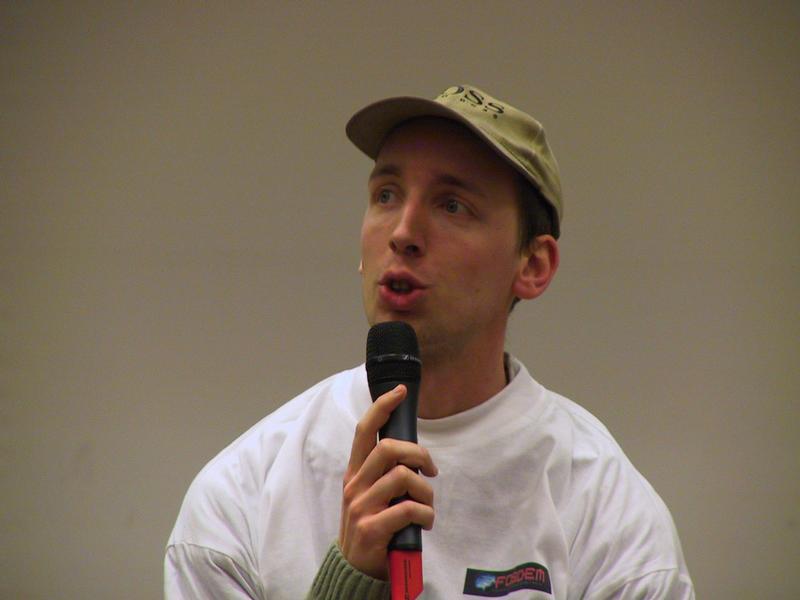 A person wearing a white Fosdem shirt and a cap speaks into a microphone during an event.