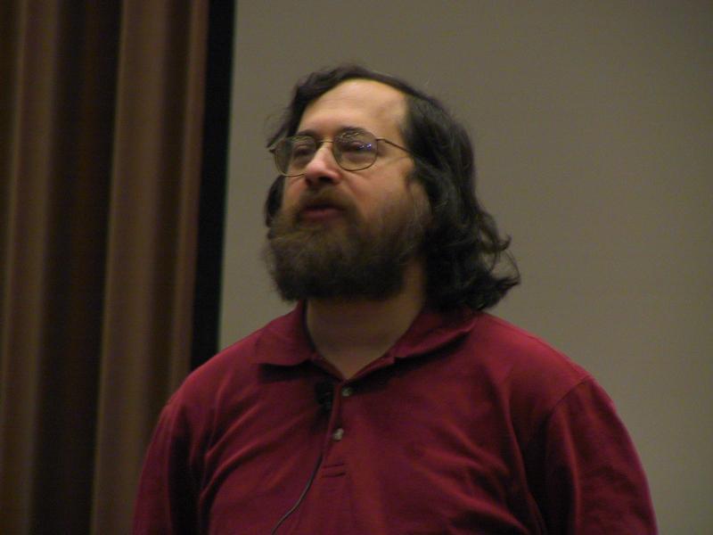 A person with glasses and a beard wearing a red shirt speaks into a microphone at an event.