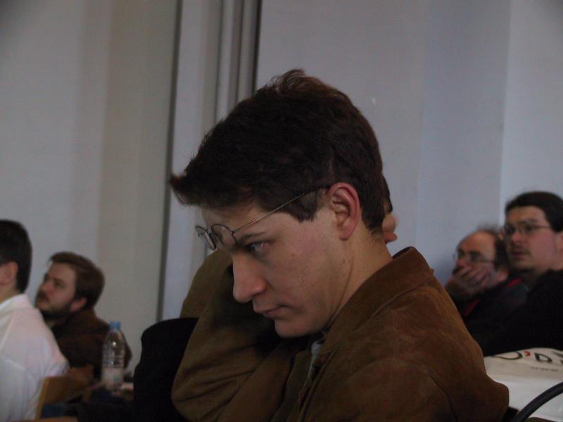 A person in a brown jacket sits in a conference room, adjusting their glasses while listening attentively.
