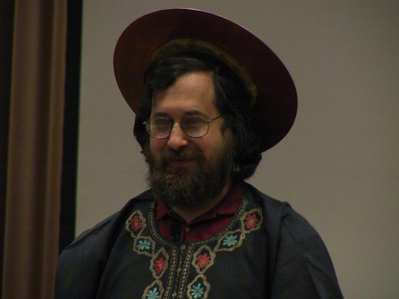 A bearded man wearing glasses and a decorated outfit stands indoors, smiling slightly.