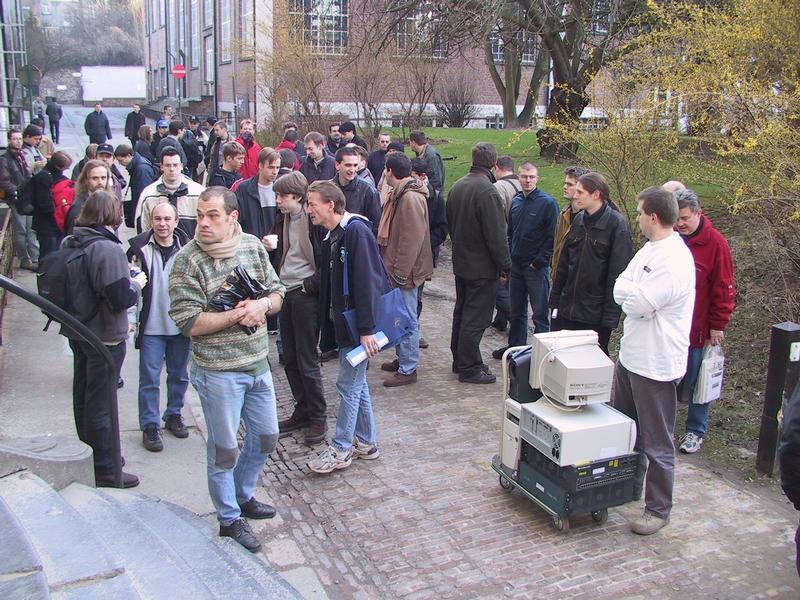 A group of people gathers outdoors, some talking and holding papers, while a cart carries old computer equipment.