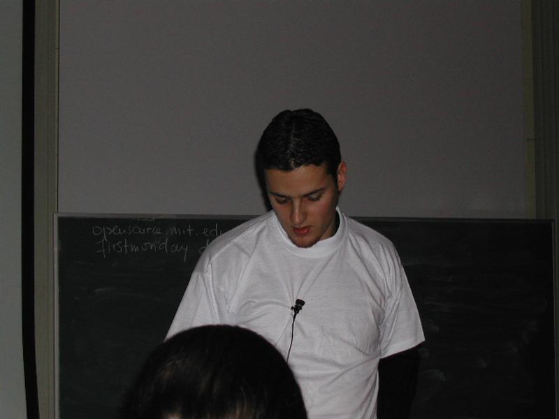 A person in a white t-shirt with a microphone speaks in front of a chalkboard at a conference.