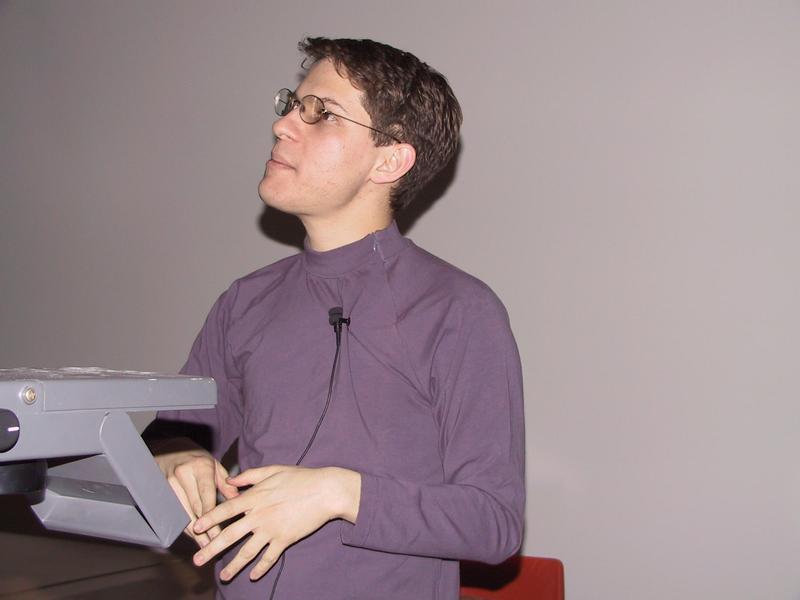 A person wearing glasses and a microphone headset stands at a podium, appearing to give a presentation.