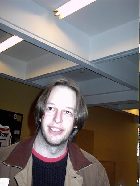 A person with shoulder-length hair and a jacket is smiling at the camera in an indoor setting.