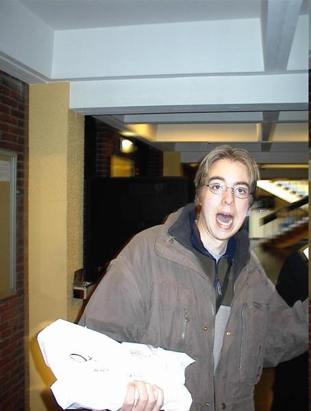 A person wearing glasses and a brown jacket holds a white bag and appears to be speaking or reacting.