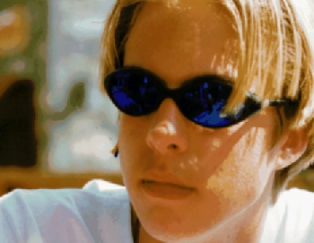 A person wearing blue sunglasses and a white shirt looks ahead with a neutral expression.