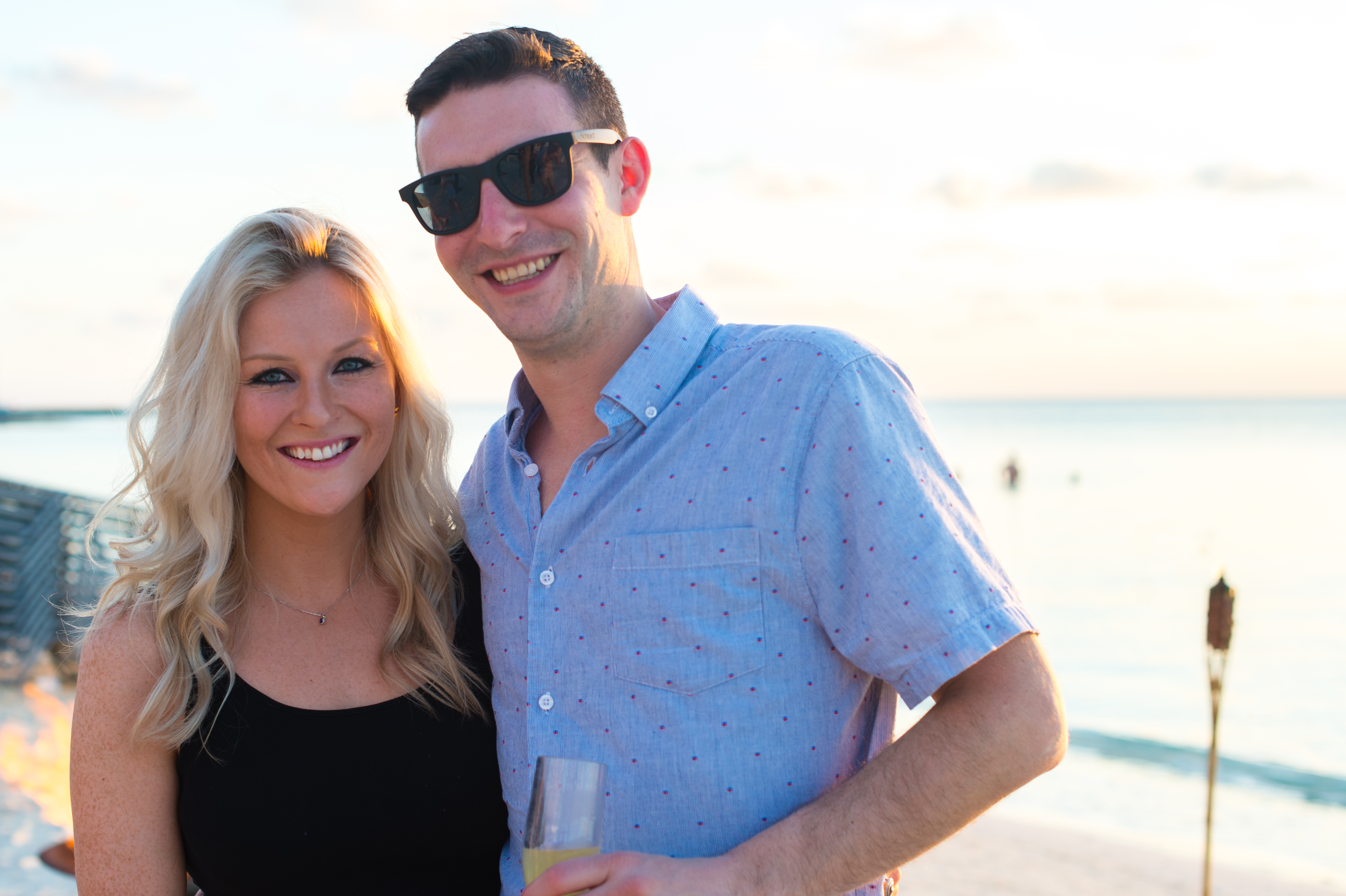 A smiling couple poses on the beach at sunset, with one holding a drink during a company trip.