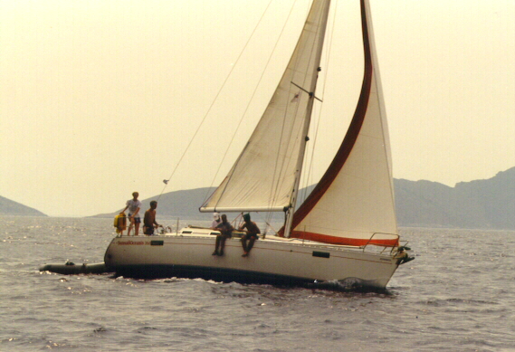 A sailboat with several people on board moves through the water near distant hills.