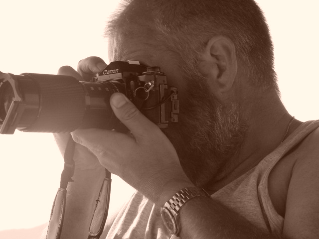 A man with a beard holds a camera with a long lens, looking through the viewfinder.