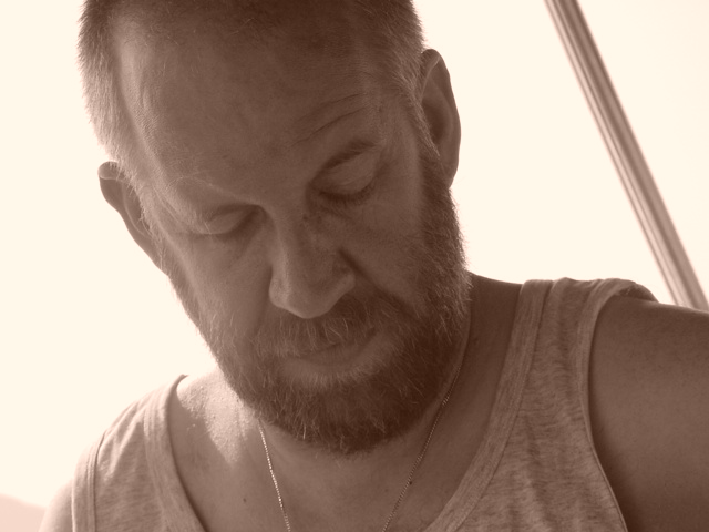 A bearded man in a tank top looks downward with a focused expression.