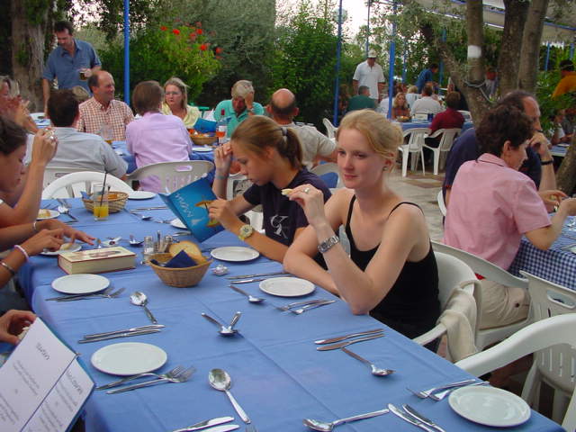 People sitting at a long table with blue tablecloths, some reading menus and others talking at an outdoor restaurant.