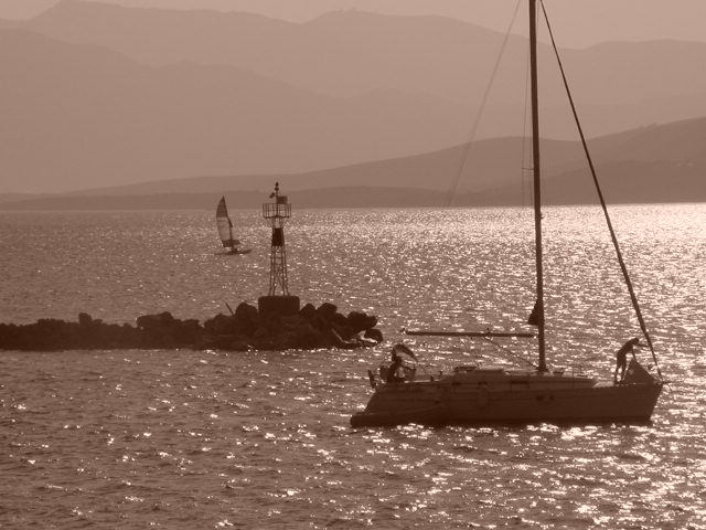 A sailboat with two people moves near a rocky breakwater with a small lighthouse, while a windsurfer passes nearby.