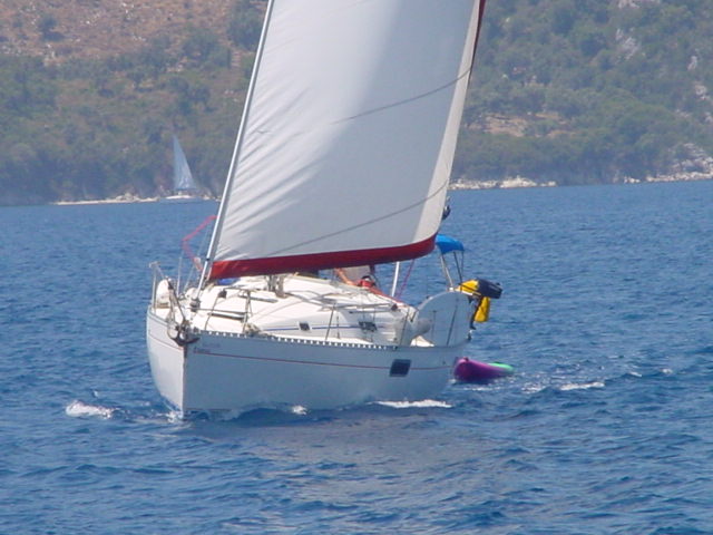 A white sailboat with raised sails moves through the blue water near a green, hilly coastline.