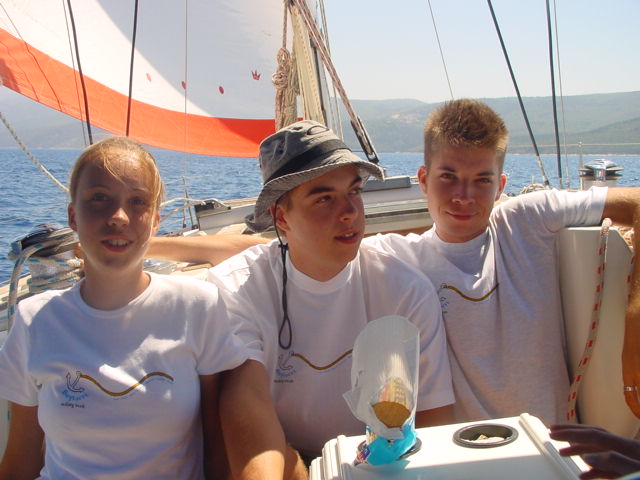 Three young people wearing matching white shirts sit on a sailboat, smiling and relaxing under the sun.