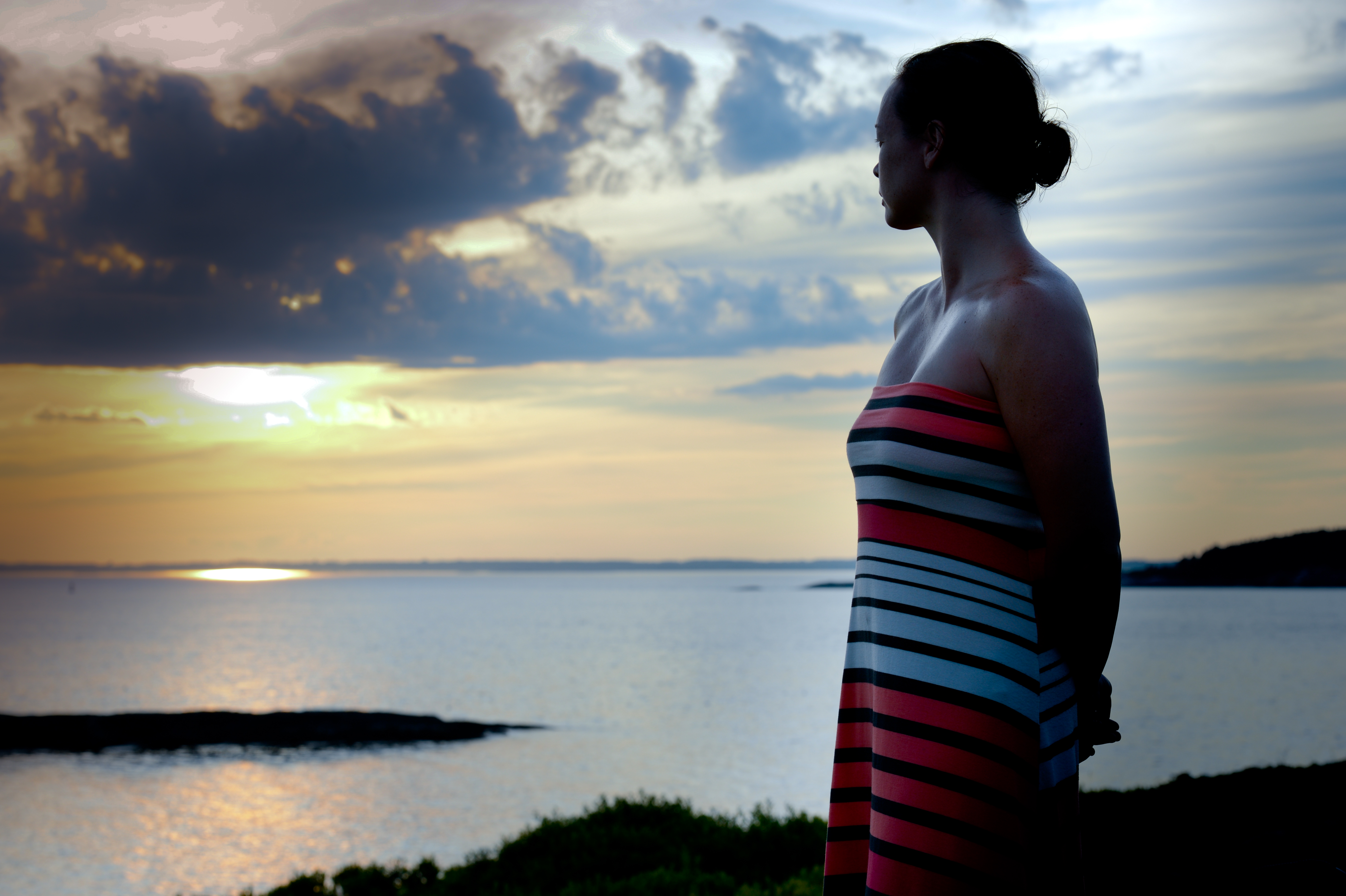 A woman in a striped dress stands near the water, looking at the sunset over the ocean.