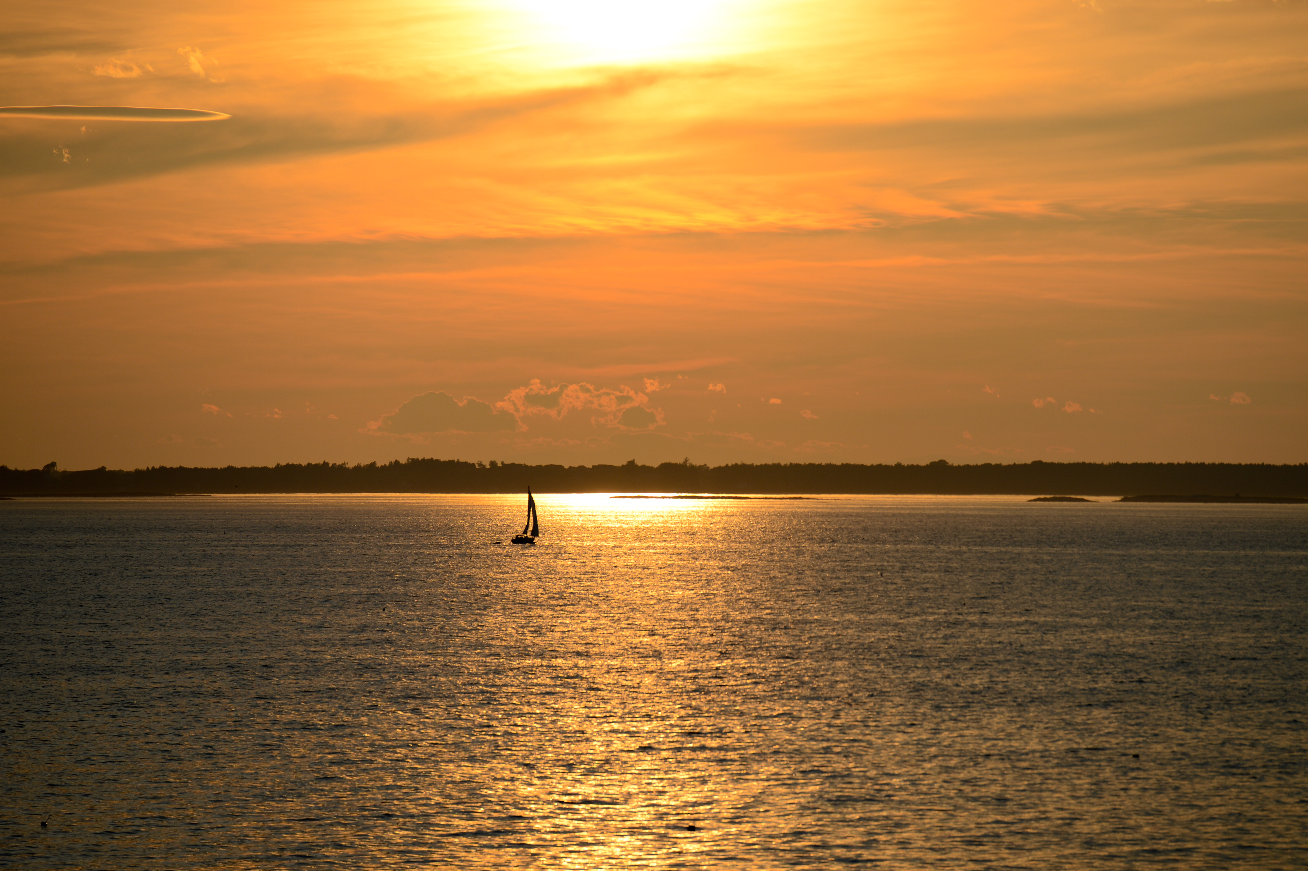 A small sailboat moves across the water under a golden sunset near the shoreline.