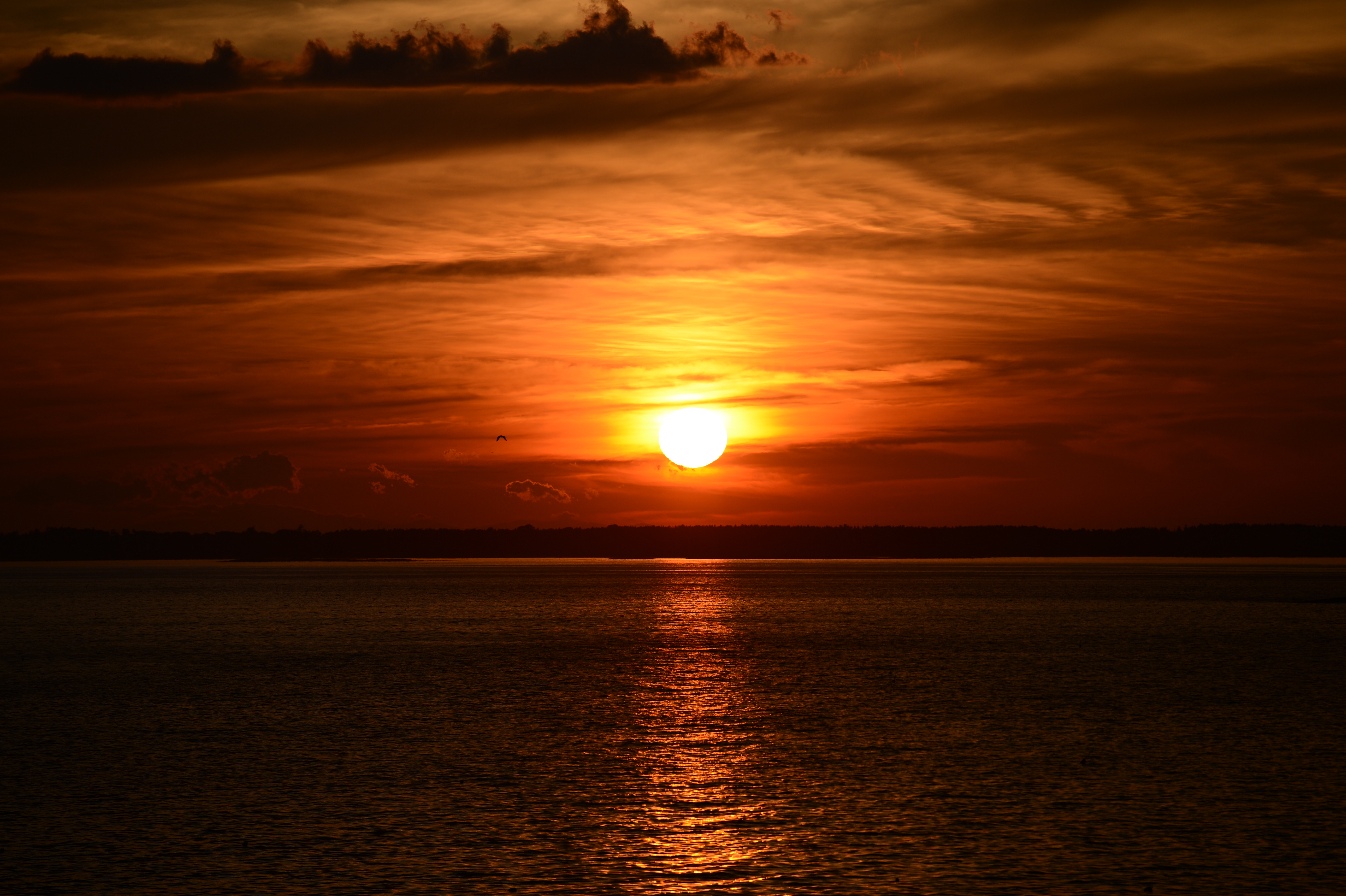 The sun sets over calm water, casting an orange glow across the sky and reflecting on the surface.