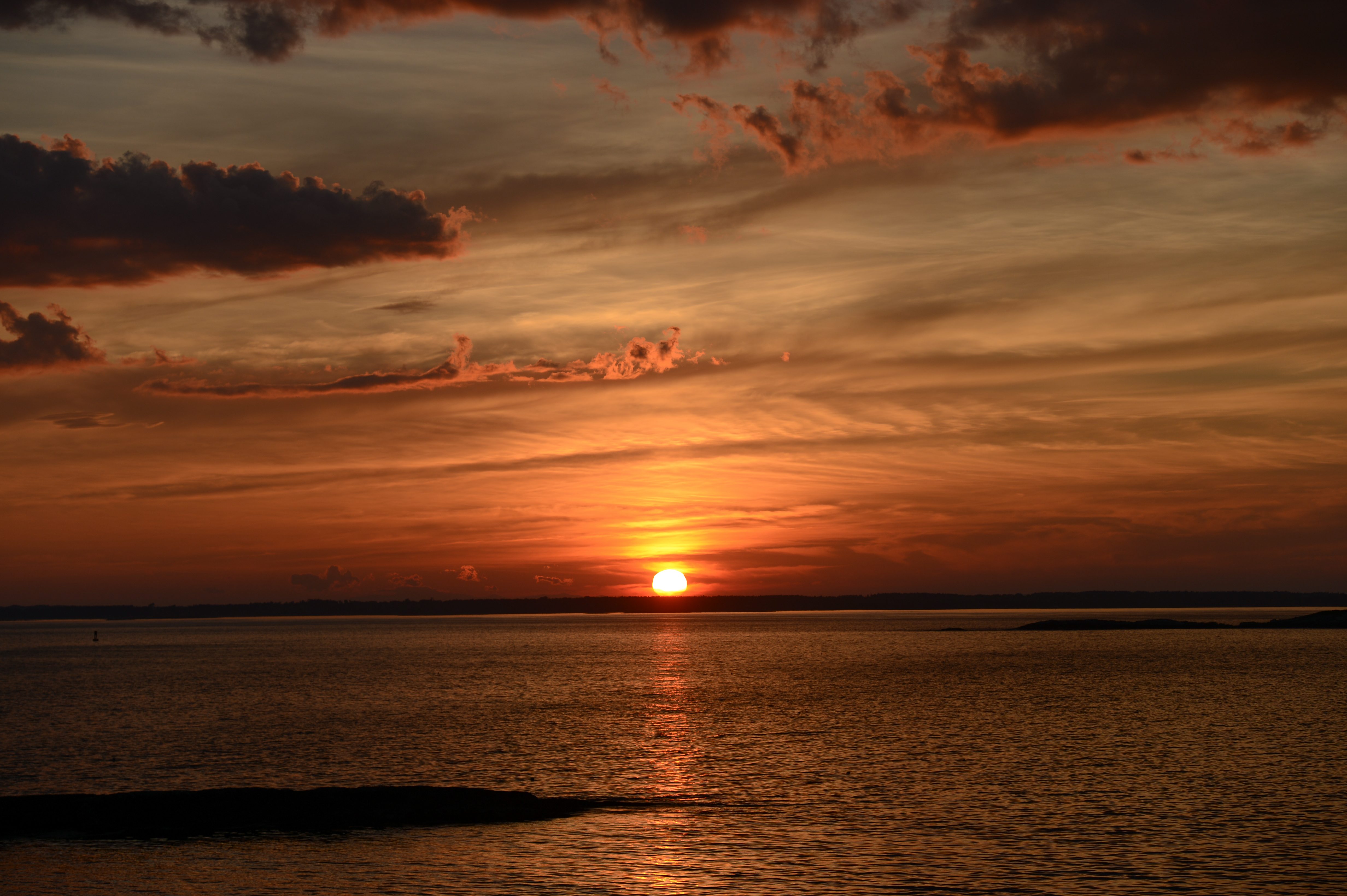 The sun sets over calm ocean waters, casting an orange glow across the sky with scattered clouds.