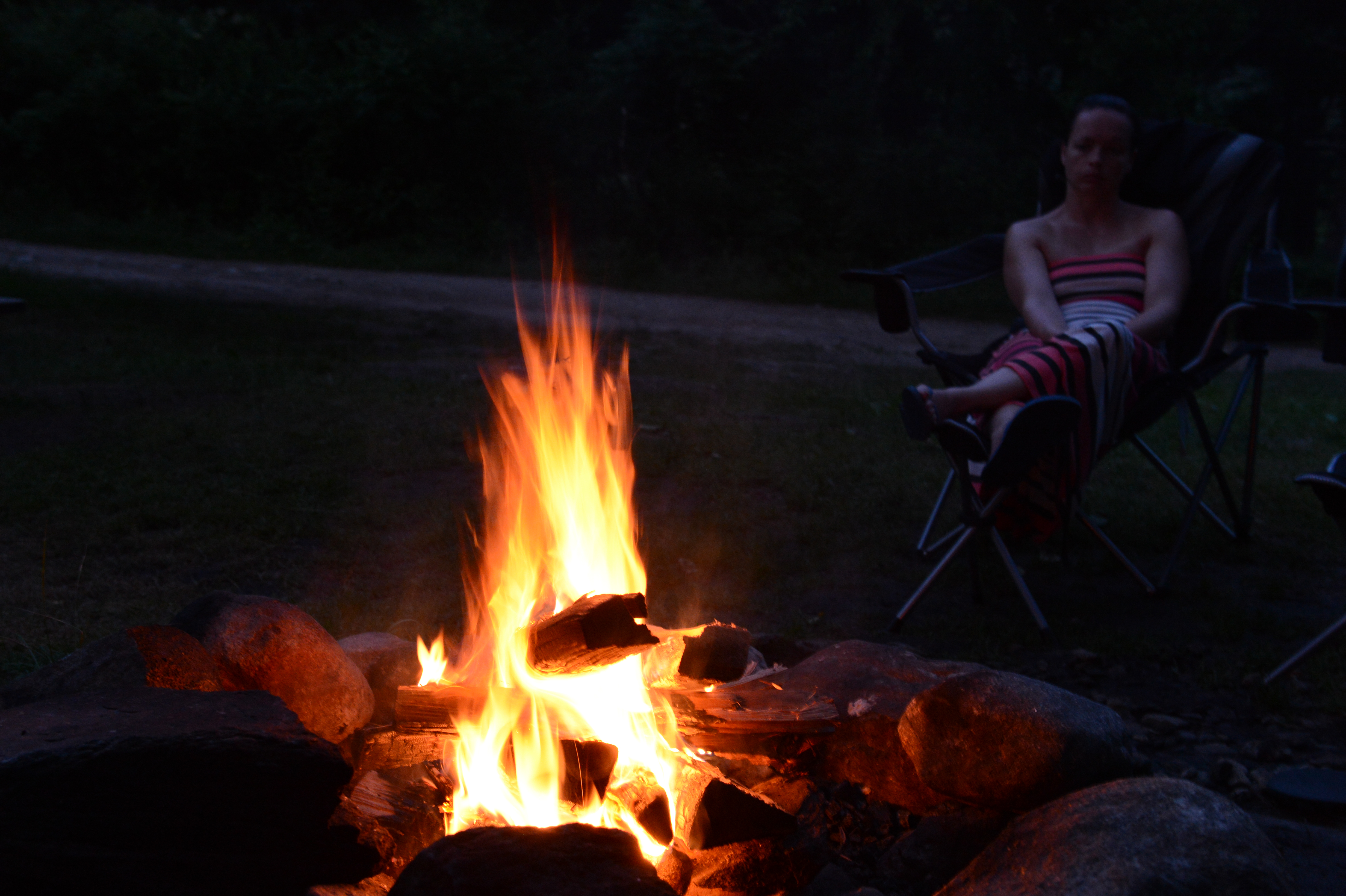 A campfire burns brightly with logs and rocks, while a person sits in a chair in the background.