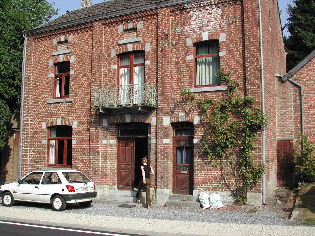 A person walks into a brick house with red window frames, while a white car is parked nearby.