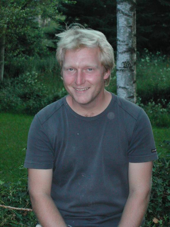 A man with short blond hair and a dark t-shirt smiles while sitting outdoors near trees and grass.