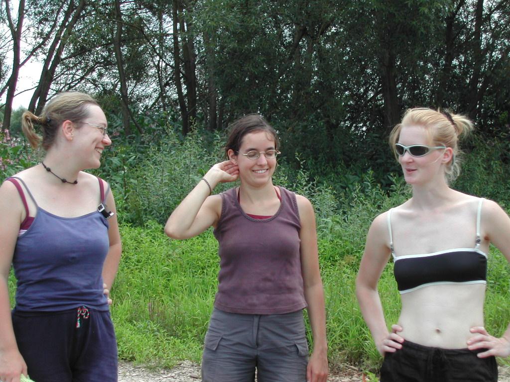 Three women stand outdoors, smiling and talking, dressed in casual summer clothing.