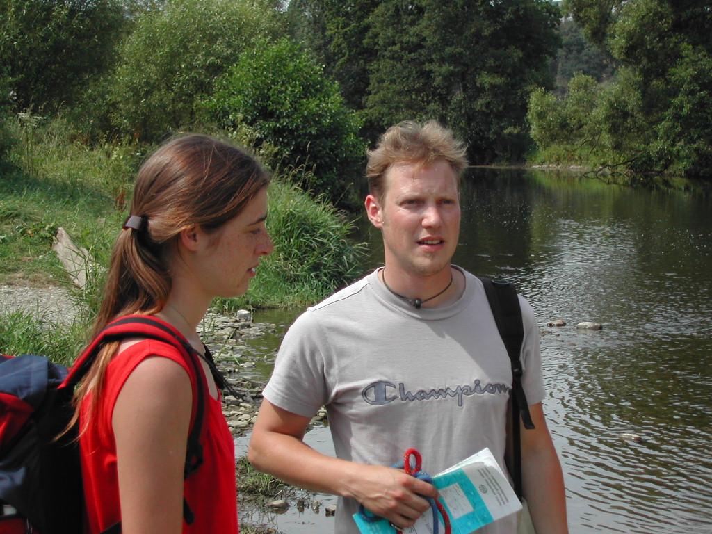 Two people with backpacks stand near a river, one holding a map and rope, appearing to discuss something.