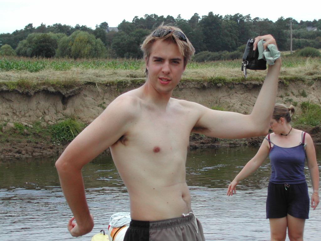 A shirtless man flexes his arm and holds a small bag near a river, while a woman stands behind him.
