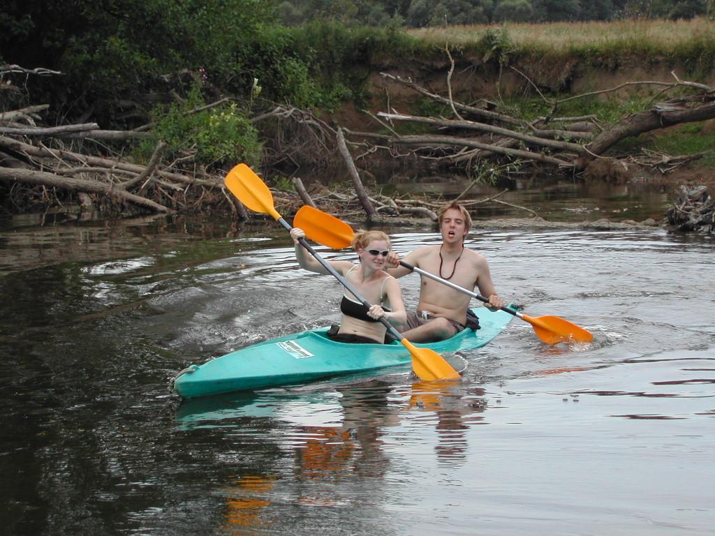 Two people paddle a green kayak on a calm river, using orange paddles to navigate through the water.
