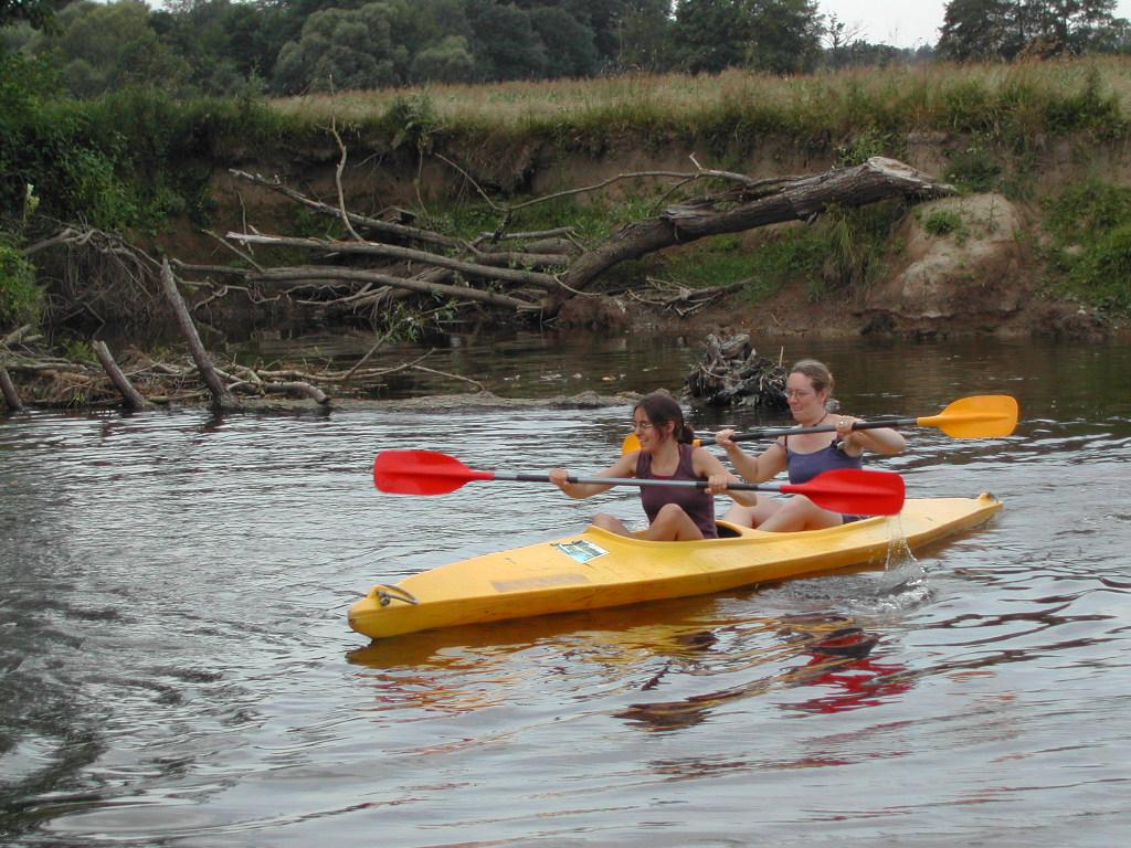Two people paddle a yellow kayak on a calm river, using red and orange paddles to move forward.