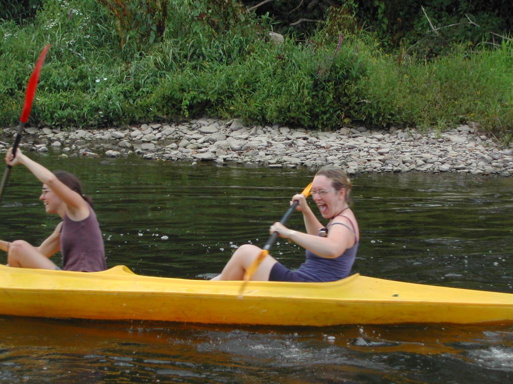 Two people paddle a yellow kayak on a river, smiling and splashing water as they move forward.